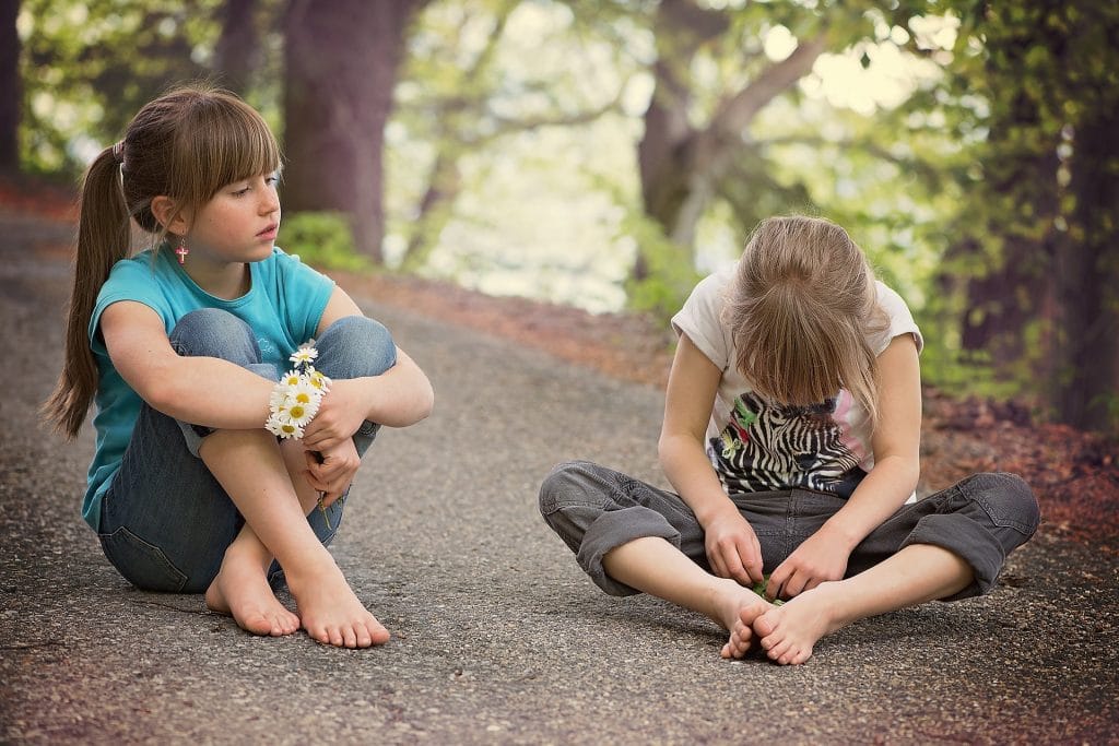 Two girls engaging in social communication, sitting on the ground in a forested area. 